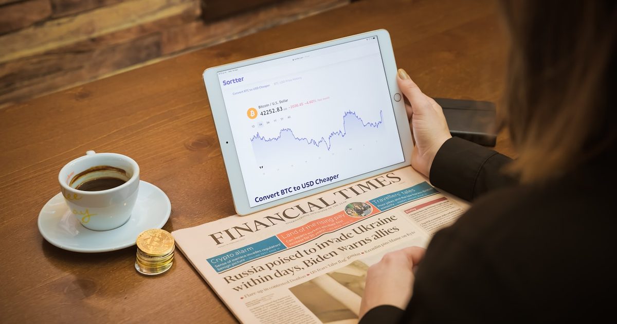 A woman reviewing market charts on a laptop alongside a financial newspaper